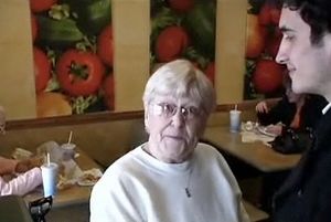 an older lady with white hair and glasses, in a subway fast food resteraunt, looking into the camera and holding Joe Hawley's hand